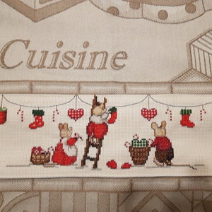 Könnte beinhalten: Kreuzstichstoff mit dem Wort "Cuisine" über einer festlichen Szene. Drei Mäuse sind dargestellt, die mit Zuckerstangen, Herzen und Strümpfen dekorieren. Geschenke und ein Korb mit Süßigkeiten sind ebenfalls vorhanden. Die Farbpalette umfasst Rot, Grün und Beige.