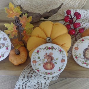Può includere: Una composizione autunnale con ricami a punto croce raffiguranti un riccio, uno scoiattolo e un gufo, circondati da ghirlande floreali. La scena include zucche, foglie autunnali e bacche, creando un'estetica calda e stagionale.