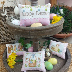 Può includere: Un vassoio di legno a due livelli con un cuscino rosa e bianco ricamato con "Buona Pasqua". Il vassoio contiene anche tre cuscini più piccoli con scene pasquali ricamate, tra cui coniglietti, pulcini e un cestino di uova. Il vassoio è decorato con narcisi gialli e uova di Pasqua pastello.