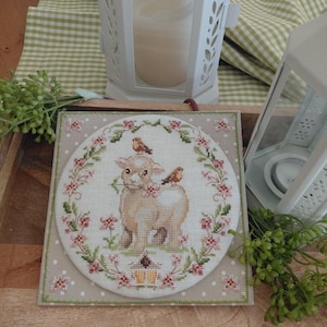 May include: A cross-stitch design featuring a lamb with two birds perched on its back, surrounded by a floral wreath. The design is on a square, dotted fabric, with a green border. The background includes a wooden tray, a white lantern, and greenery.