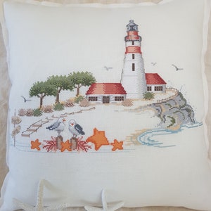 Può includere: Un faro bianco e rosso con un tetto rosso si trova su una scogliera rocciosa che domina un mare blu. Due gabbiani sono appollaiati su un palo di legno in primo piano, e ci sono stelle marine e coralli sparsi sulla spiaggia.