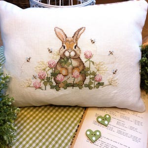 Può includere: Un cuscino bianco con un ricamo a punto croce di un coniglio marrone seduto in un campo di fiori rosa e bianchi con api che volano intorno. Il cuscino è su una superficie a scacchi verde e bianca.