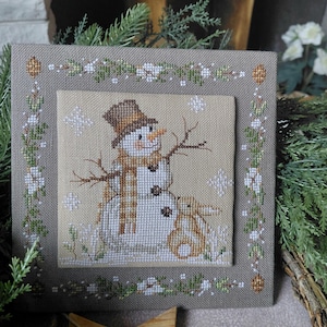 Peut inclure: Broderie au point de croix représentant un bonhomme de neige et un lapin. Le bonhomme de neige porte un chapeau et une écharpe bruns, avec des bras en brindilles. Le lapin est beige. L'œuvre est encadrée d'une bordure florale et de flocons de neige, créant un thème hivernal.