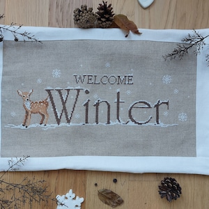 Può includere: Un ricamo a punto croce su lino beige con un bordo bianco. Il ricamo presenta un cervo marrone e il testo "Welcome Winter".