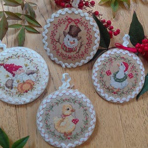 Może przedstawiać: Cztery okrągłe, haftowane ozdoby świąteczne z uroczymi wzorami. Jeden przedstawia bałwana, drugi kaczątko z sercem, trzeci gnoma, a ostatni gęś. Każdy ma białe koronkowe wykończenie i pętelkę do zawieszenia.