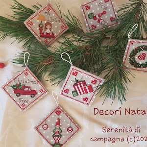 Może przedstawiać: Pięć ręcznie robionych ozdób choinkowych z czerwonymi i zielonymi haftami na beżowym tle. Ozdoby przedstawiają choinkę, wieniec, lalkę, prezent i gnoma. Ozdoby są oznaczone napisami "Christmas Tree", "My Dolly", "Toys", "Decor Natalizi" i "Serenita di campagna (c) 2020".
