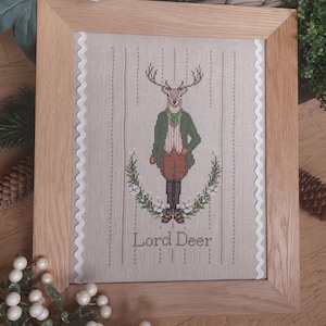 May include: A framed cross-stitch embroidery featuring a deer wearing a green jacket and a bow tie. The deer is standing in front of a wreath of flowers and the text "Lord Deer" is below the deer.