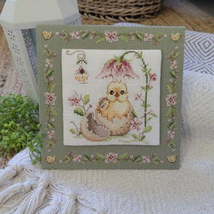 May include: A framed cross-stitch featuring a yellow chick hatching from an egg, surrounded by pink flowers and greenery. The artwork is set against a light green background with a floral border, evoking a springtime theme.