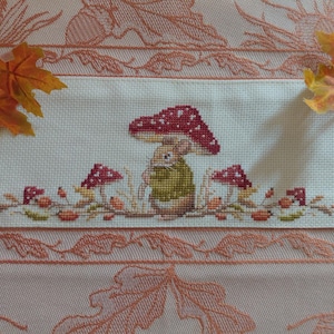 Pode incluir: Um bordado em ponto cruz sobre tecido branco apresentando um rato castanho com uma camisola verde sob um grande cogumelo vermelho com pintas brancas. O design inclui cogumelos mais pequenos e detalhes de folhagem de outono em tons de vermelho e castanho.