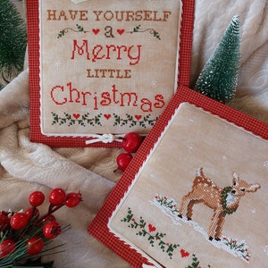 May include: Two framed cross-stitch pieces with a red and white checkered border. The top piece says "Have Yourself a Merry Little Christmas". The bottom piece features a brown deer with a green wreath around its neck standing in the snow.