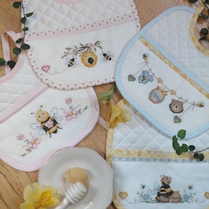 Puede incluir: Una colección de baberos acolchados para bebés con diseños bordados de abejas y miel. Los baberos presentan ribetes rosas, azules y amarillos, con ilustraciones de punto de cruz de abejas, panales y tarros de miel. También hay un plato blanco pequeño con miel y un cucharón.