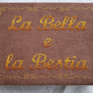 Può includere: Un oggetto in tessuto marrone con la scritta "La Bella e la Bestia" ricamata con filo giallo e arancione. Il testo è centrato e incorniciato da un bordo decorativo.
