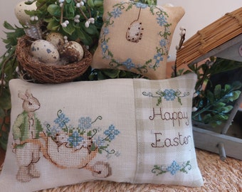 May include: Two embroidered pillow covers with an Easter theme. One pillow features a bunny carrying a wheelbarrow full of blue flowers and the other pillow says "Happy Easter" with blue flowers on either side.