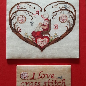 Può includere: Un ricamo a punto croce che raffigura una donna seduta su una sedia, circondata da una cornice a forma di cuore di rami e fiori. Il ricamo include le lettere A e B e il testo "Amo il punto croce".