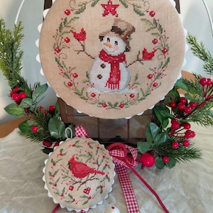 Può includere: Un ricamo a punto croce circolare raffigurante un pupazzo di neve con sciarpa e cappello rossi, circondato da una ghirlanda di rami di pino verdi e bacche rosse. Un ricamo circolare più piccolo mostra un uccello cardinale rosso su un ramo.