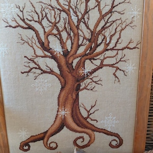 May include: Cross-stitch artwork of a bare tree with brown branches and trunk, set against a beige background. White snowflake accents are scattered throughout the design. The artwork is framed in a wooden frame.