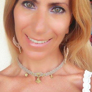 May include: A woman wearing a beige macrame choker necklace with gold accents and a white top. The necklace has a large, ornate pendant in the center.