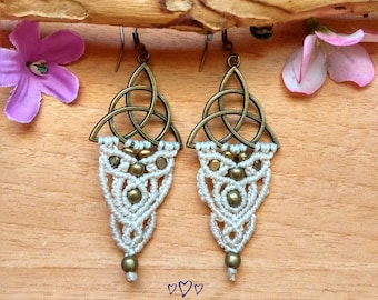 May include: White macrame earrings with gold beads and a bronze Celtic knot charm. The earrings are hanging from a wooden surface with pink and purple flowers.