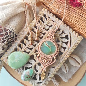 Puede incluir: Un collar de macramé hecho a mano con un gran colgante ovalado de piedra verde claro. El collar presenta un cordón beige con cuentas doradas y un pequeño dije dorado. Se exhibe sobre una superficie de madera tallada con elementos naturales.