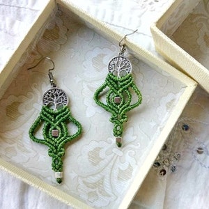Puede incluir: Un par de pendientes de macramé verde con dijes de árbol de la vida plateados. Los pendientes se muestran en una caja blanca con un patrón floral.