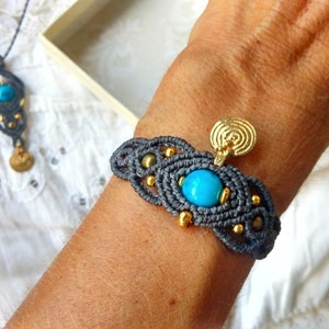 Könnte beinhalten: Graues Makramee-Armband mit einem türkisfarbenen Stein und einem goldenen Anhänger. Das Armband befindet sich am Handgelenk einer Person. Eine passende Halskette mit einem türkisfarbenen Stein und einem goldenen Anhänger befindet sich im Hintergrund.