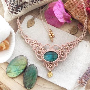Puede incluir: Un collar de macramé rosa con una gran piedra ovalada de labradorita verde y detalles dorados. El collar se muestra sobre una superficie de tela blanca con dos piedras de labradorita más pequeñas.