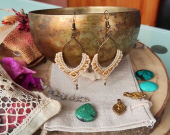May include: A pair of handmade macrame earrings with a cream and gold color scheme. The earrings are made with a delicate, intricate knotting technique and feature a small, dangling bead at the bottom.
