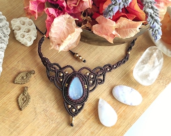 Puede incluir: Un collar gargantilla de macramé marrón con un colgante de piedra de luna azul y cuentas doradas. El collar se muestra sobre una superficie de madera con varias piedras blancas pequeñas.