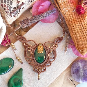 Puede incluir: Un collar con un colgante de piedra de malaquita verde con un acento de metal dorado, tejido con cordón marrón y cuentas doradas. El collar se muestra sobre una superficie de tela blanca con otras piedras de malaquita verde y una piedra de amatista púrpura.