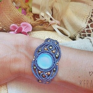 Puede incluir: Un brazalete de macramé azul con una piedra de ópalo azul claro y cuentas doradas. El brazalete está en la muñeca de una persona.
