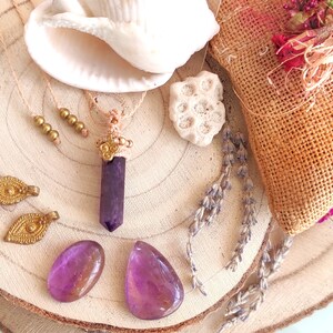 Puede incluir: Un collar con un colgante de cristal de amatista púrpura con detalles dorados y un cordón de macramé. El collar se muestra sobre una superficie de madera con una concha, un pequeño trozo de coral y lavanda seca.