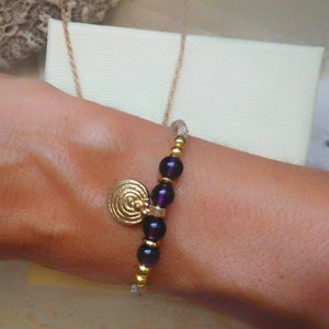 May include: A beaded bracelet featuring a gold spiral charm, dark purple beads, and gold accents. The bracelet is strung on a neutral-colored cord. A gold chain and a cream-colored box are in the background.