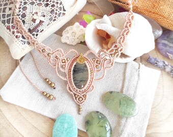 Puede incluir: Un collar de macramé hecho a mano en color melocotón suave, con un colgante de piedra ovalado con un patrón gris oscuro. El collar está adornado con pequeñas cuentas doradas y un intrincado trabajo de nudos, mostrando un estilo bohemio.