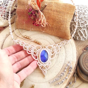 Puede incluir: Un collar de macramé blanco con un colgante de piedra azul y cuentas doradas. El collar se muestra sobre una superficie de madera.