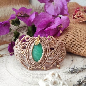 May include: A light brown macrame bracelet with a green gemstone and gold accents. The bracelet is on a wooden surface with flowers and a burlap bag in the background.