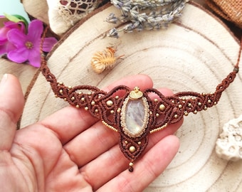 Puede incluir: Un collar de macramé marrón con un cierre de metal dorado y un colgante de piedra preciosa transparente e iridiscente.