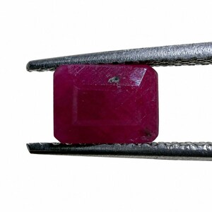 May include: A rectangular, deep red ruby gemstone with a faceted surface and a small dark inclusion near the top edge, held securely between the textured metal tips of a pair of silver-toned tweezers against a plain white background.