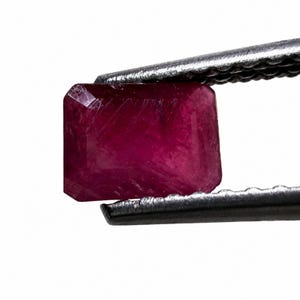 May include: A rectangular faceted ruby gemstone with a deep red color and visible internal inclusions, held securely by a pair of silver-toned metal tweezers against a plain white background. The stone displays a polished surface with sharp edges.