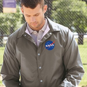 May include: A gray, button-up jacket with a NASA logo on the left chest. The logo is a blue circle with a white outline and a red rocket.