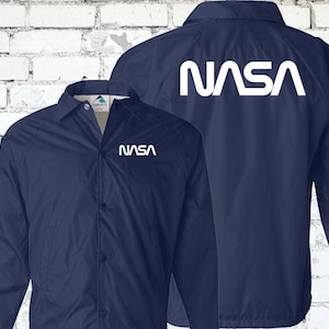 Puede incluir: Una chaqueta azul marino con un logotipo blanco de la NASA en la parte posterior y delantera. La chaqueta tiene cierre de botones y mangas largas.