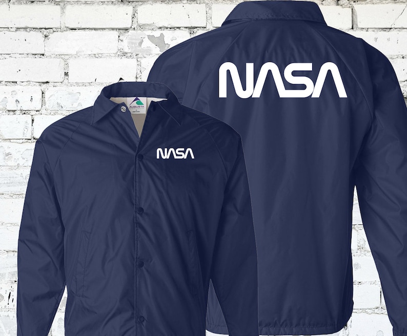 Puede incluir: Una chaqueta azul marino con un logotipo blanco de la NASA en la parte posterior y delantera. La chaqueta tiene cierre de botones y mangas largas.