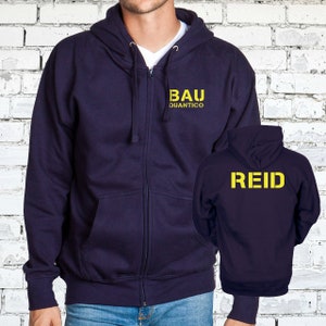 May include: A navy blue zip-up hoodie with a yellow embroidered logo that says "BAU Quantico" on the front and "REID" on the back.