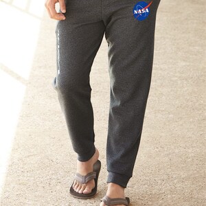 May include: Gray sweatpants with a blue NASA logo on the left leg. The sweatpants have a cuffed bottom and are worn with brown flip-flops.
