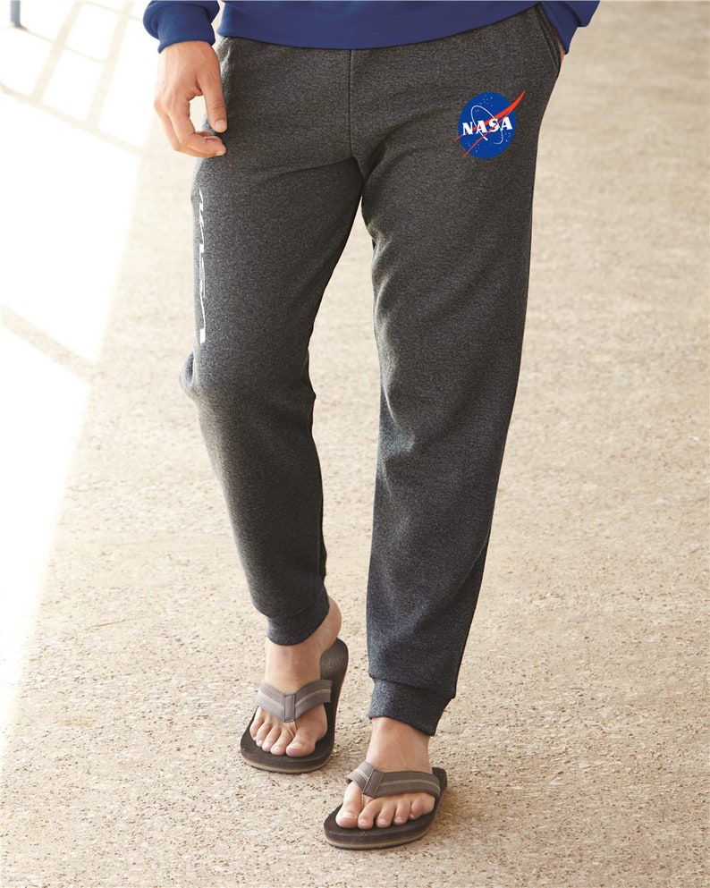 May include: Gray sweatpants with a blue NASA logo on the left leg. The sweatpants have a cuffed bottom and are worn with brown flip-flops.
