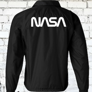 Puede incluir: Chaqueta cortavientos negra con un logotipo blanco de la NASA en la espalda.