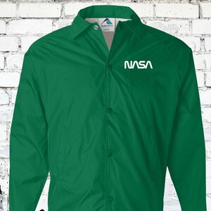 Puede incluir: Una chaqueta verde con botones y un logotipo blanco de la NASA en el pecho izquierdo.