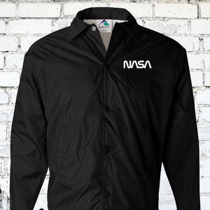 Puede incluir: Una chaqueta de nylon negra con un logotipo blanco de la NASA en el pecho izquierdo. La chaqueta tiene un cierre de botones en la parte delantera y un cuello de pie.