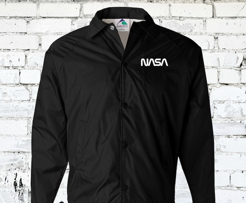 Puede incluir: Una chaqueta de nylon negra con un logotipo blanco de la NASA en el pecho izquierdo. La chaqueta tiene un cierre de botones en la parte delantera y un cuello de pie.