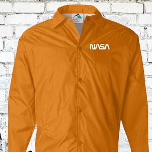 Puede incluir: Una chaqueta de nailon naranja brillante con cierre de presi&oacute;n blanco y un logotipo "NASA" blanco en el pecho izquierdo.