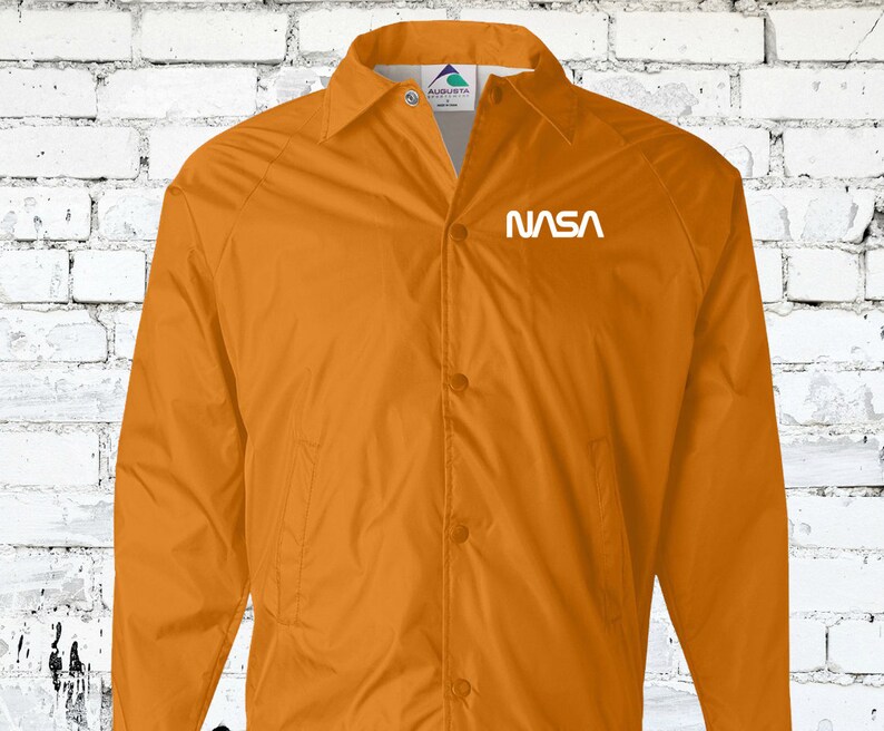 Puede incluir: Una chaqueta de nailon naranja brillante con cierre de presi&oacute;n blanco y un logotipo "NASA" blanco en el pecho izquierdo.